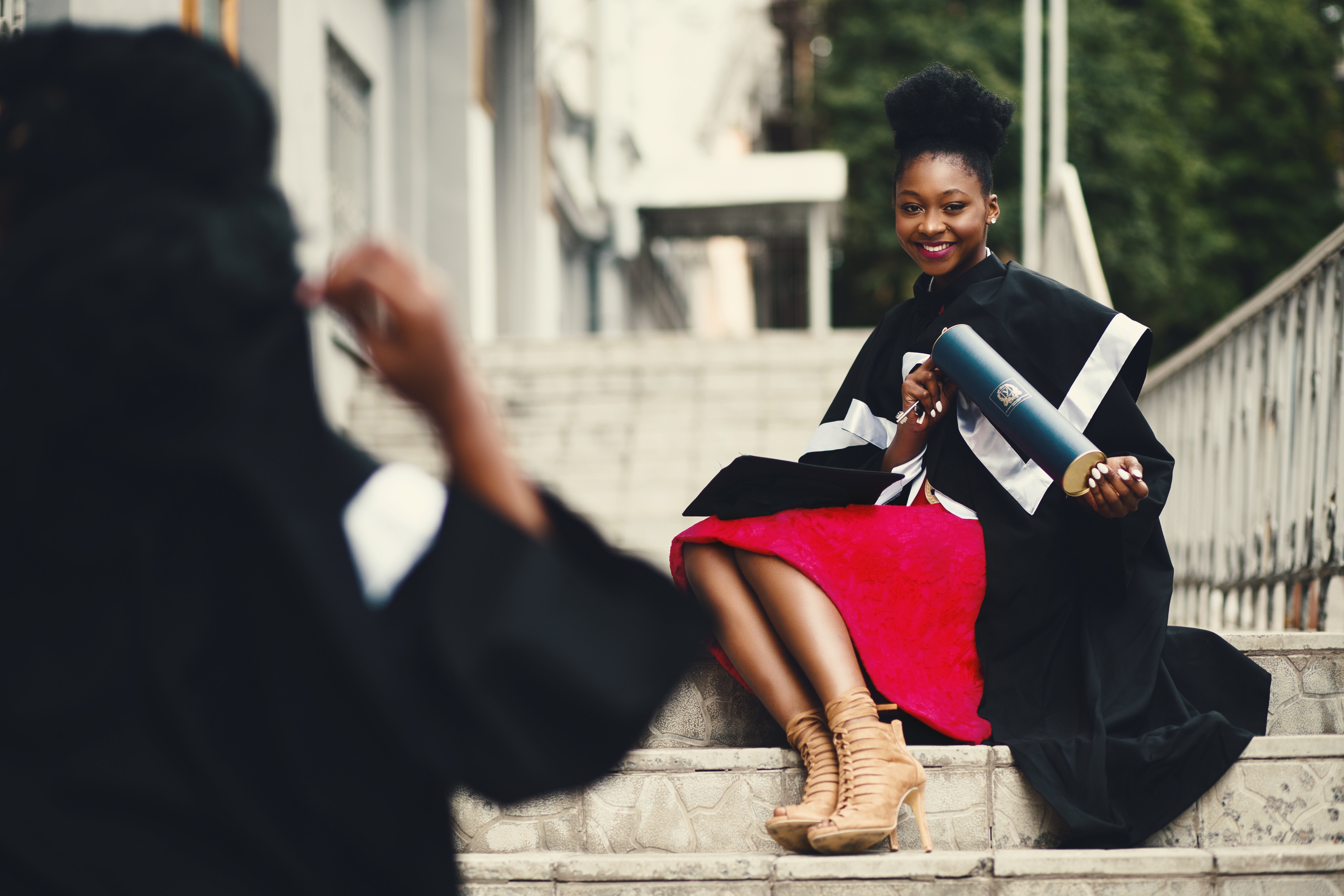 A young Black woman sitting on the steps of a building posing with a college degree.